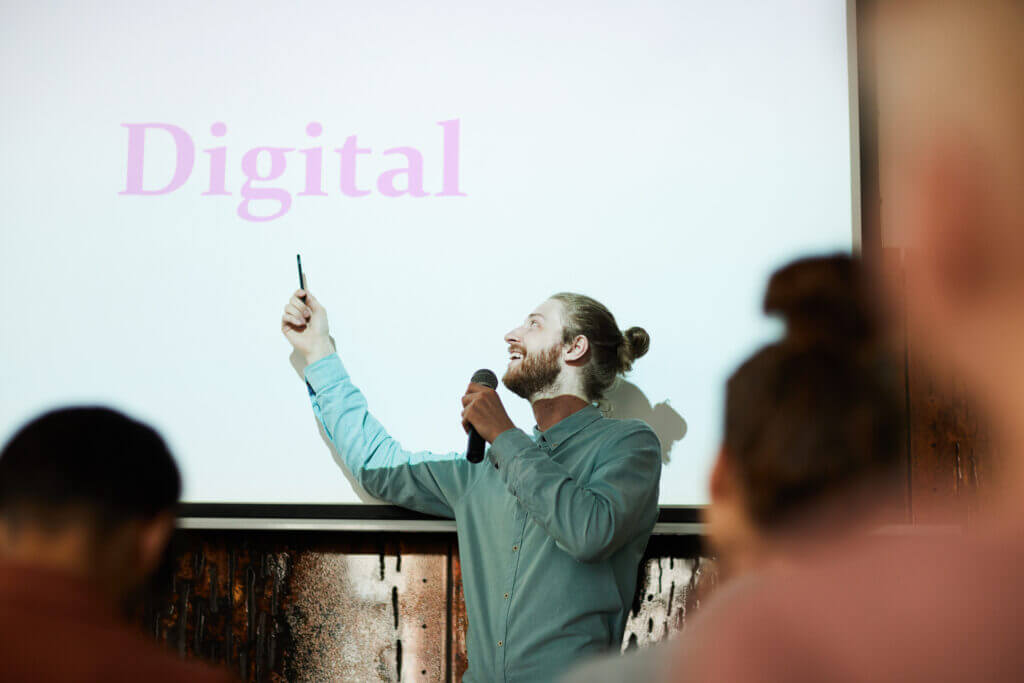Waist up portrait of contemporary bearded man giving presentation pointing at word DIGITAL on projector screen during lecture, copy space